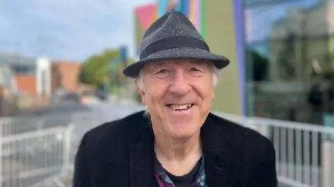BBC/George Carden Ken Pollack smiles into the camera. He wears a grey trilby, a black jacket and a black t-shirt.