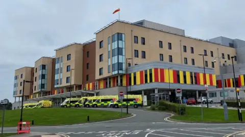 A hospital with ambulances parked outside it. The hospital building is several storeys high and red, black and yellow outside on the lower part.