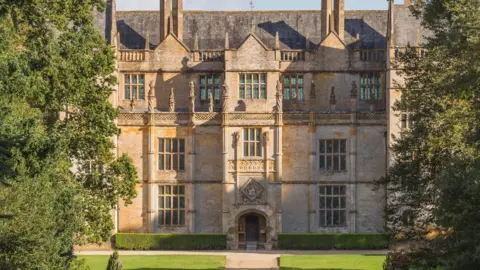 National Trust A view of the front of Montacute House, a mansion with a grand entrance and big windows. There are two trees in the foreground of the photo.