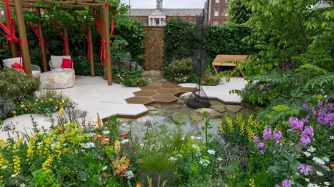 RHS/Neil Hepworth A picturesque garden seen with colourful flowers blooming. There is a seated area in the corner with red ribbons hanging from it.