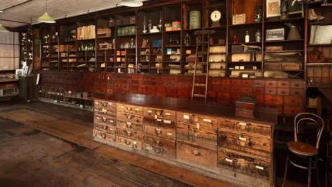 Historic England The large shop interior has wooden shelving and drawers across its back wall, a wooden counter with large drawers and a wooden chair to the side.