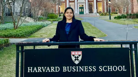 Shreya Mishra Reddy stands behind a sign with the words Harvard Business School and a crest of the institution at its campus in Boston in the United States.