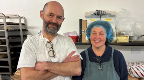 A man and woman wearing aprons smile at the camera - they are in a bakery and the woman wears a hair net.