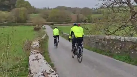 Three men cycling down a country lane. They are wearing yellow hi-vis and helmets.