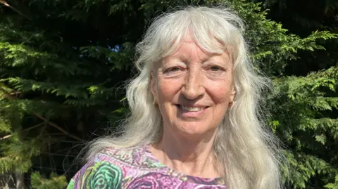 Jenny Connor has long grey hair and a fringe and is wearing a top with purple and green fowers on it, smiling at the camera while standing in front of some hedgerows on a sunny day.