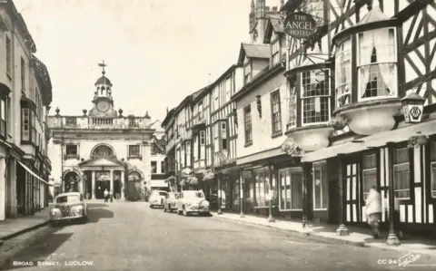 Walter Scott A black and white photograph of a street with black and white wooden buildings on the right side and a building with a large pillared entrance at the end of the street.