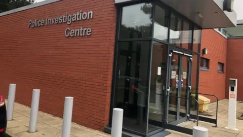 Vikki Irwin/BBC This is a picture of the Martlesham police investigation centre. On one side of the building, in grey letters against red bricks, it spells out  police investigation centre . There are automatic sliding doors at the front of the building  and a free standing intercom visible. There are also white bollards running alongside the building. 