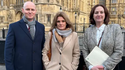 Ellen Roome stands between Max Wilkinson, MP for Cheltenham, and Lola McEvoy, MP for Darlington, outside the Houses of Parliament. They are wearing coats.