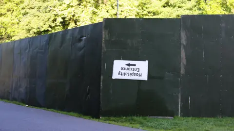 Emma Gibbs A hospitality entrance sign hangs upside down on a green metal fence in front of foliage in Heaton Park