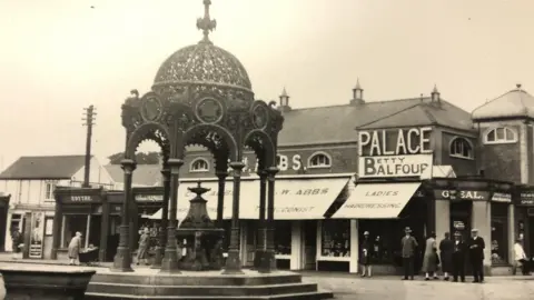 March and District Museum Black and white picture of the Coronation Fountain from 1929, complete with water drinking fountain in the centre. Smartly dressed men and women can be seen near shops in the background, wearing hats suits and coats.