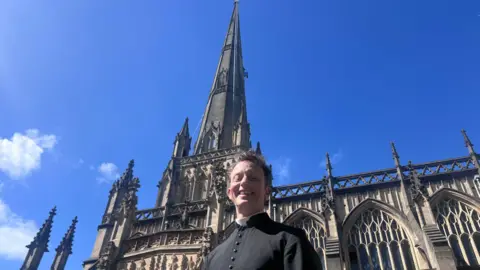 A vicar with short grey hair wearing black clerical clothing is standing in front of the St Mary Redcliffe church spire on a sunny day. 