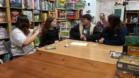 A group of adults and children sit around talking, behind them is a wall of shelves filled with board games.