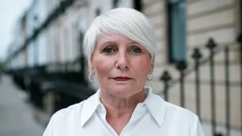 GMB Scotland A woman with white hair, large silver earrings and wearing a white blouse, staring at the camera