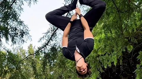 A man hangs upside from a rope with his legs apart at the knees. He's looking down. Behind him are trees and a blue sky.