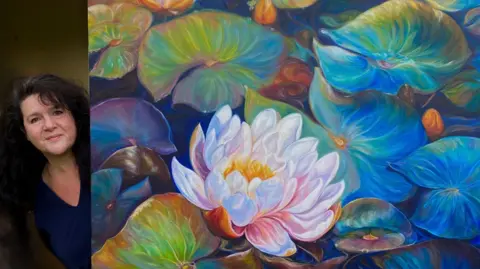 A dark haired woman next to a floral painting with bright colours.