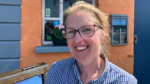 Sarah Cochrane stands in front of a building which has an orange-painted wall with a white-framed window. She is smiling and wearing glasses, with blond hair pulled back behind her head anda blue and white checked shirt.