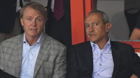 Nassef Sawiris in a blue shirt and dark jacket sits to the left of fellow Aston Villa co-owner Wes Edens, who is wearing a light checked jacket and light blue shirt. 