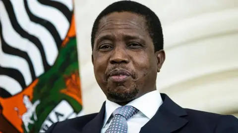 Former Zambian President Edgar Lungu siting down in a suit and tie with the Zambian flag behind him