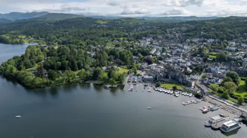 An aerial view of Bowness-on-Windermere, a lake shore town in the Lake District National Park. There are lots of boats moored in the marina and the town stretches out beyond it, surrounded by trees. In the distance are hills and mountains.