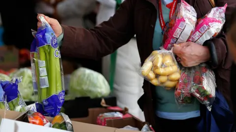 A person picking up a pack of celery with their right hand while holding other groceries including a pack of potatoes in their other hand. The person is wearing a turquoise jumper and brown coat