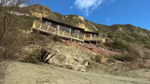 Two dark wooden beach chalets on a cliffside in Branscombe where they have become uneven and the decking has partially collapsed.