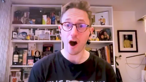 A man in a black T-shirt and glasses looks at the camera with his mouth open in surprise