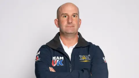 Royal British Legion A bald white man in a navy blue branded jacket and a white shirt stands in front of a blank backdrop.