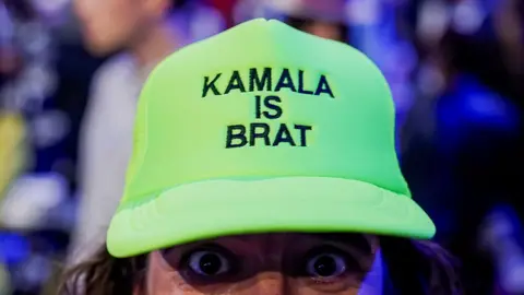 EPA A Kamala Harris supporter wears a green 'Kamala is Brat' hat with the eyes of the person peeking out from under the cap.