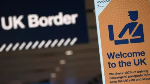 A picture of the UK border. A sign which says UK border can be seen in the background. In front of it is a yellow sign with blue writing which says " Welcome to the UK". 
