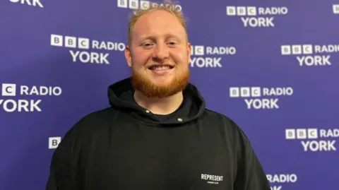 The image shows Cain North standing in front of a blue BBC Radio York backdrop. He is wearing a black hooded sweatshirt with the word “REPRESENT” printed in white on the left side of the chest. 