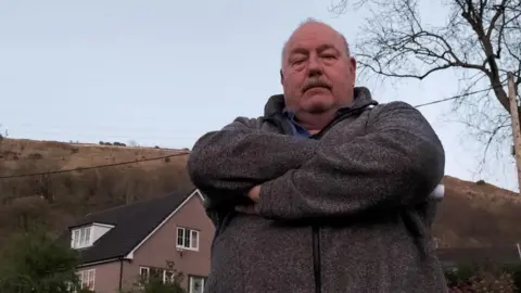 LDRS A bald man with a moustache wearing a grey fleece, looking angry with his arms crossed across his body. Behind him are houses and hills.