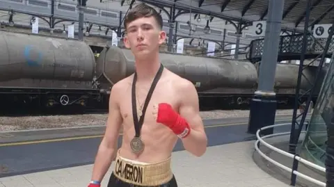 Supplied Cameron Laidlaw is standing on a railway station platform with a grey freight train on the tracks behind him. To one side is a grey, painted column with a white, square sign sticking out from it showing this is platform three. Cameron has short, brown hair and a serious expression. He is not wearing a top and has on black boxing trousers with a gold ruched waistband with his name on it in black letters. He has red binding around his hands and wrists and is wearing a medal on a black ribbon and what looks like a smaller gold medallion necklace.