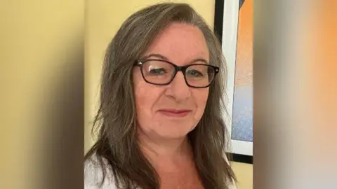 Shropshire Amateur Swimming Association A woman with long brown hair and dark framed glasses in front of a yellow wall and a white-framed window