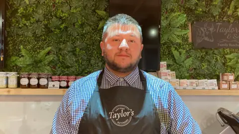 A man with short dark hair, a checked shirt and black butcher's apron standing behind the counter in his shop.