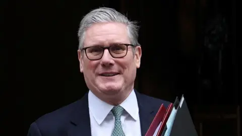 Reuters Sir Keir Starmer in front of the 10 Downing Street door. He is wearing a dark coloured suit, white shirt, green tie and holding a red folder and a black folder.