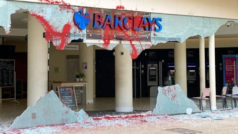 Smashed window at the entrance of Barclays Bank with red paint sprayed around building