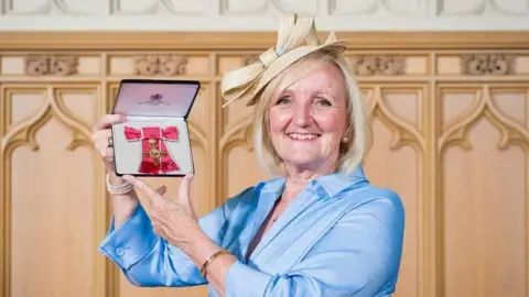 Supplied Sandra a woman, wearing a blue dress and a white bow hat holding a medal and smiling for camera.