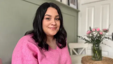 A woman with dark hair wearing a pink jumper sits at a white table, with pink and white flowers in a vase. There's a sage green wall behind her.