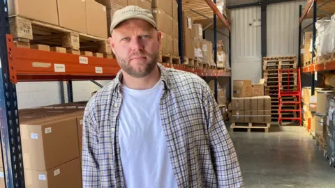A man wearing an open chequered shirt, and a white t-shirt, is standing inside a warehouse.