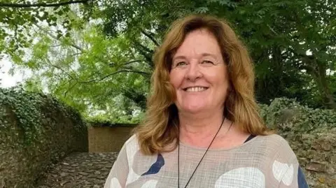 Dawn Johnson A woman smiling at the camera standing in front of a stone wall and surrounded by trees