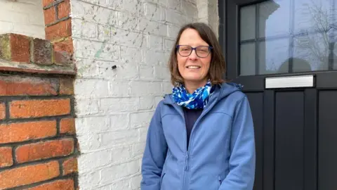A woman in a blue coat is standing in front of her house. The door is black and there is brickwork to the left.