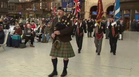 A piper wearing a kilt leads three flag bearers and a procession of veterans through Glasgow Central Railway Station, watched by passengers waiting for trains, seated on benches. 