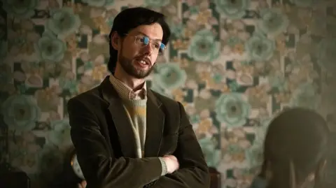 Rob Youngston/FX A man with long, dark hair and a beard, with his arms folded, wearing a velvet green blazer, metal glasses, knitted striped jumper and shirt... Pictured against 1970s style wallpaper with large floral pattern.