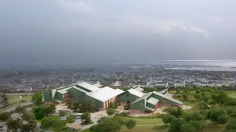 Inital designs of Peterhead community campus. The building has multiple light green sloped roofs and red/pink brick walls. It is surrounded by trees and grass, with Peterhead and the North Sea behind it. 