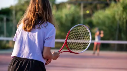 Anonymous young females playing tennis. One at the front is wearing a white t-shirt and holding a red and white racket, the other is blurred out across the net. 
