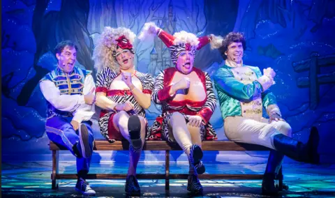 Pamela Raith Four men sit on a bench on a stage. Two are dressed in blue waist coats, the two in the middle are dressed as pantomime dames with red dresses, stockings and white wigs.