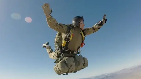 Rob during a parachute jump over the desert. His arms are stretched out, with his legs in the air behind him. He is wearing a khaki vest and a helmet with a clear visor, with what appears to be a kit bag attached to his harness. The ground below him is flat, with a mountain ridge in the distance.
