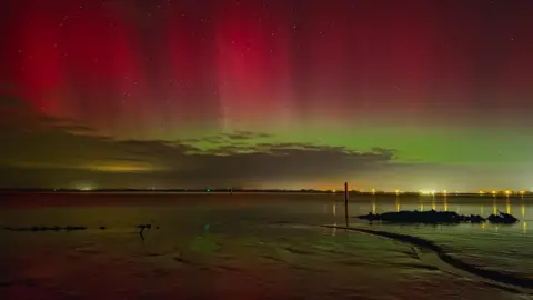 Mud flats at Great Yarmouth in shadow with yellow, green and pink skies of the Northern Lights. Lights on the edge of the shoreline are in the distance.