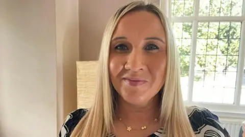Katie is wearing a patterned top and is stood by a window in a light, bright room inside a house. She has long straight blonde hair.