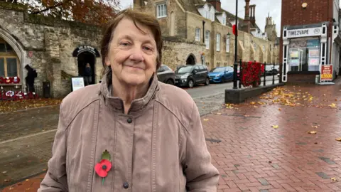 TOM JACKSON/BBC Patricia Canham is standing to the left of the photo and smiling at the camera. She is wearing a light-coloured jacket with a red paper poppy affixed on the left-hand side. Behind her is a sandstone-coloured wall with a war memorial in an alcove including the figure of a black metal solider with his head bowed and several poppy wreaths. There are also poppies adorning a nearby railing and one attached to a lamppost. 
